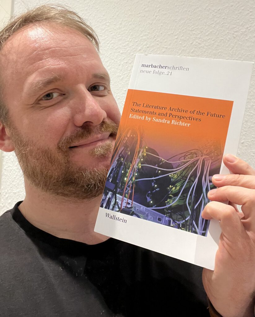 This picture shows the smiling face of Sebastian Möring holding the book the book "The literature archive of the future: statements and perspectives", edited by Sandra Richter and published in 2023 by Wallstein.