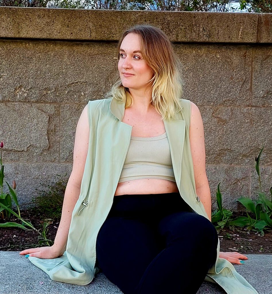 Portrait photo of Joleen Blom sitting on a concrete ledge. She is wearing pastel green and dark clothing and her gaze is directed to the left. The background shows a warm grey stone wall.