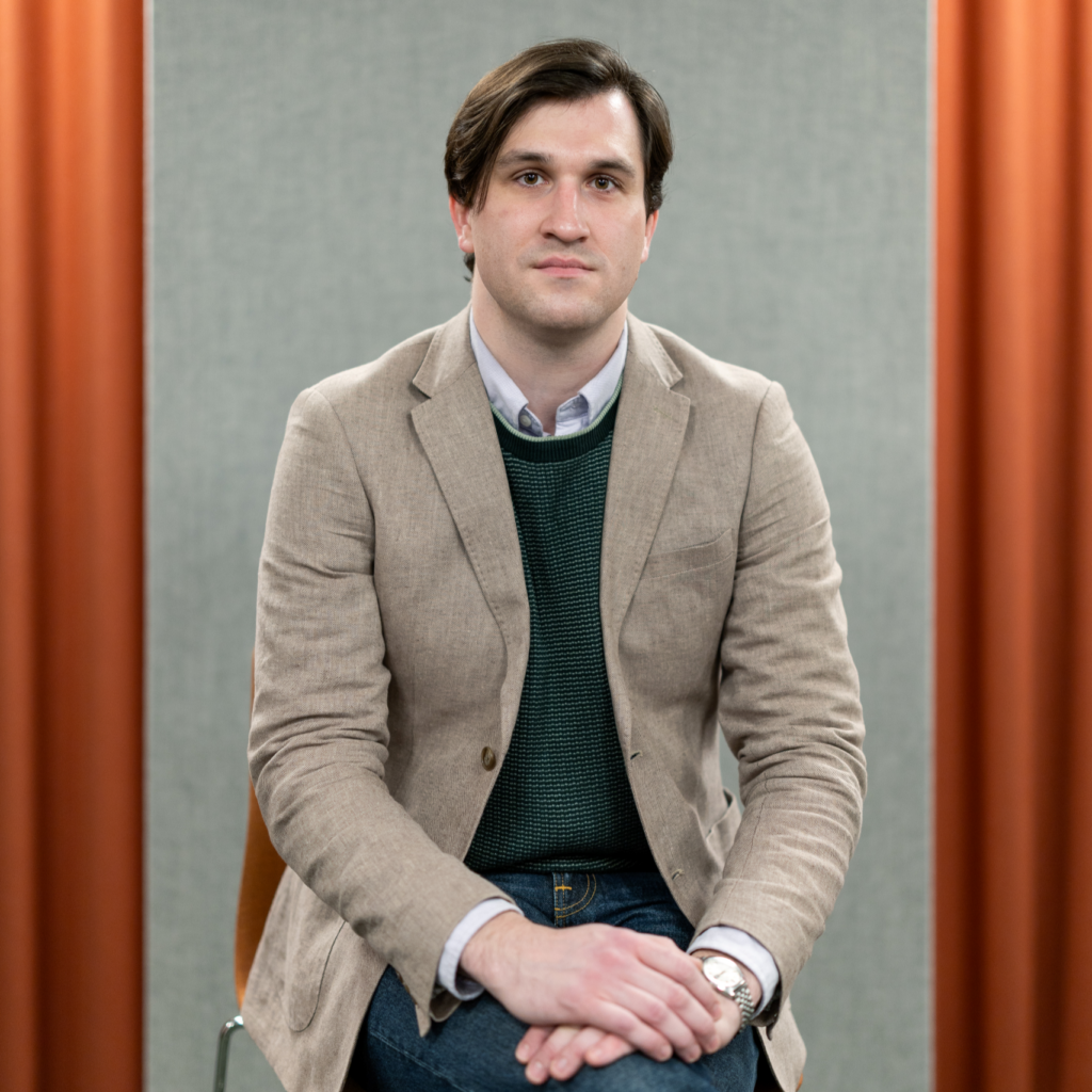 Studio portrait of a man with brown hair seated and looking directly at the camera, hands clasped in his lap. He wears a beige blazer over a dark green textured sweater and a light blue button-down shirt, with a silver wristwatch visible. The background features a gray panel flanked by warm orange curtains.