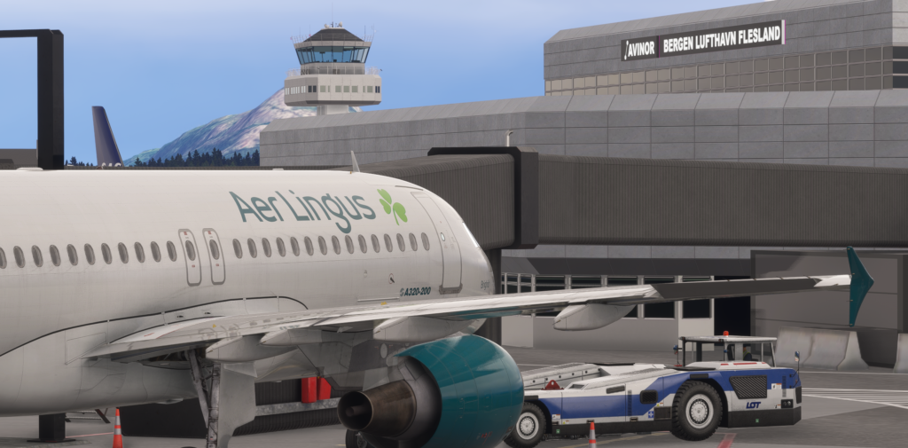 In-game screenshot from Microsoft Flight Simulator showing an Aer Lingus Airbus A320-200 parked at the gate at Bergen Airport Flesland (ENBR), Norway. The aircraft's white fuselage with green shamrock livery and teal engine nacelle dominates the foreground. A pushback tractor marked "LOT" is positioned beneath the aircraft. In the background, the terminal building displays the "Avinor Bergen Lufthavn Flesland" signage, with the airport control tower and a snow-dusted mountain visible behind it under a clear blue sky.