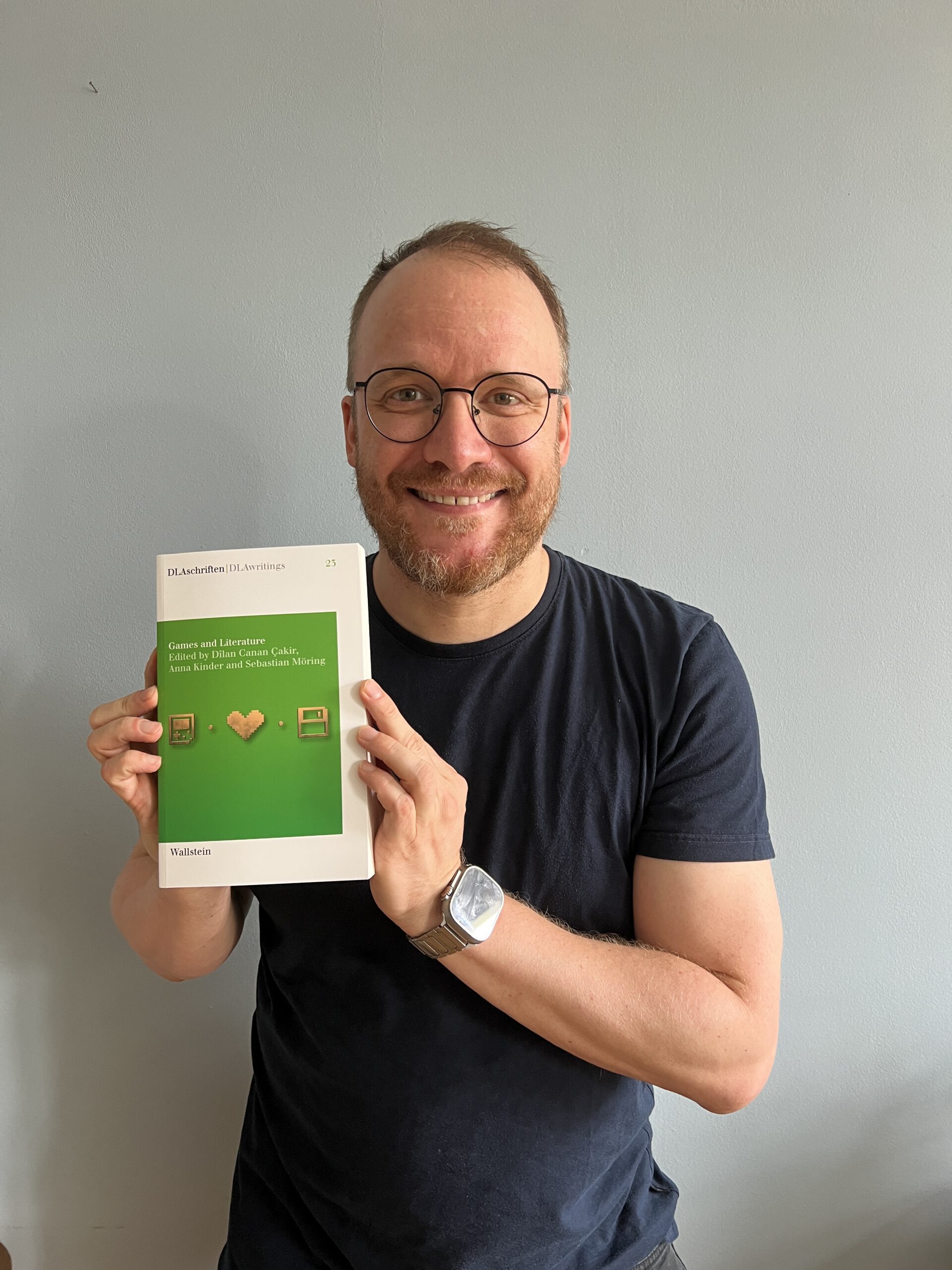 A smiling man with short brown hair, a beard, and round glasses holds up a copy of the book "Games and Literature" (edited by Dîlan Canan Çakir, Anna Kinder, and Sebastian Möring, Wallstein). He wears a dark navy T-shirt and a smartwatch, standing against a plain light grey wall. The book's white-and-green cover with three pixel-art icons faces the camera.